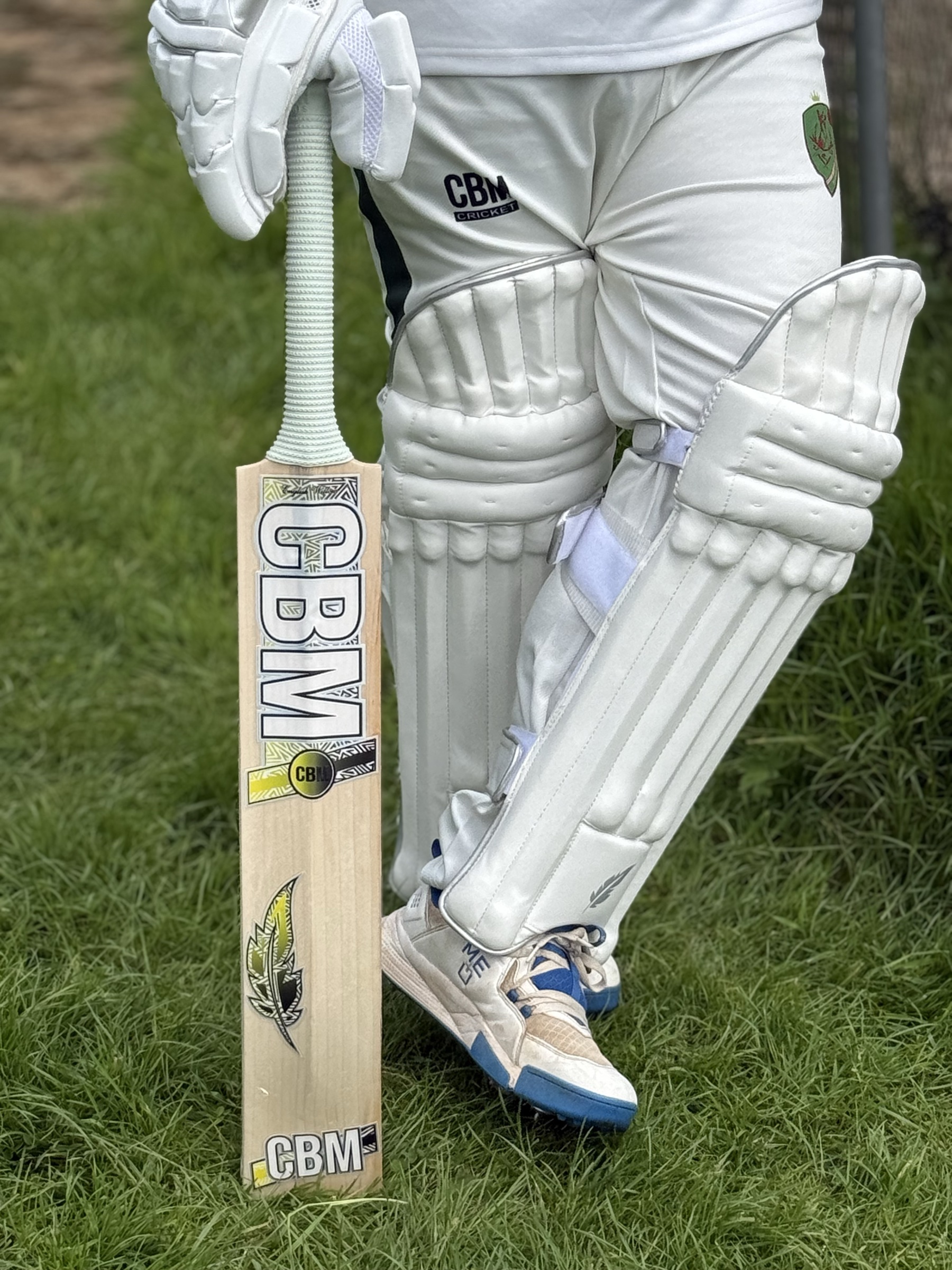 A player in CBM kit, batting pads, gloves, and a CBM bat, standing on grass.
